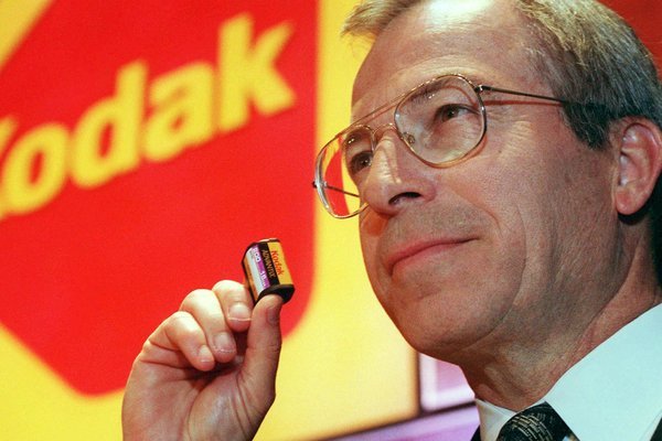 George Fisher 手持 Kodak 产品站在 Kodak 标志前，他是 Kodak 历史上第一位从外部聘请的 CEO